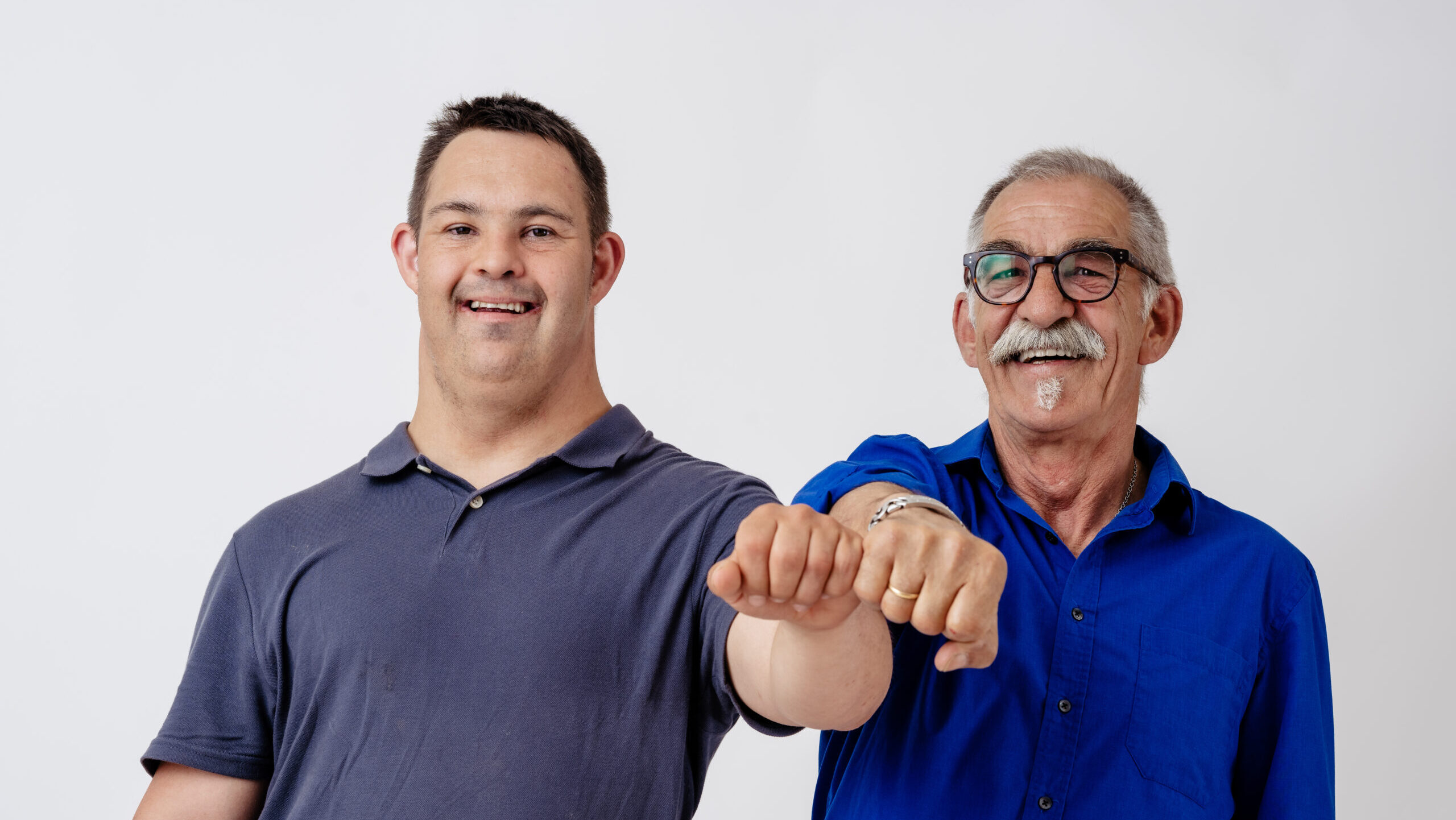 Zwei Männer stehen nebeneinander vor hellem Hintergrund und lächeln in die Kamera. Sie strecken ihre Fäuste nach vorne und berühren sich freundschaftlich mit einem „Fist Bump“.