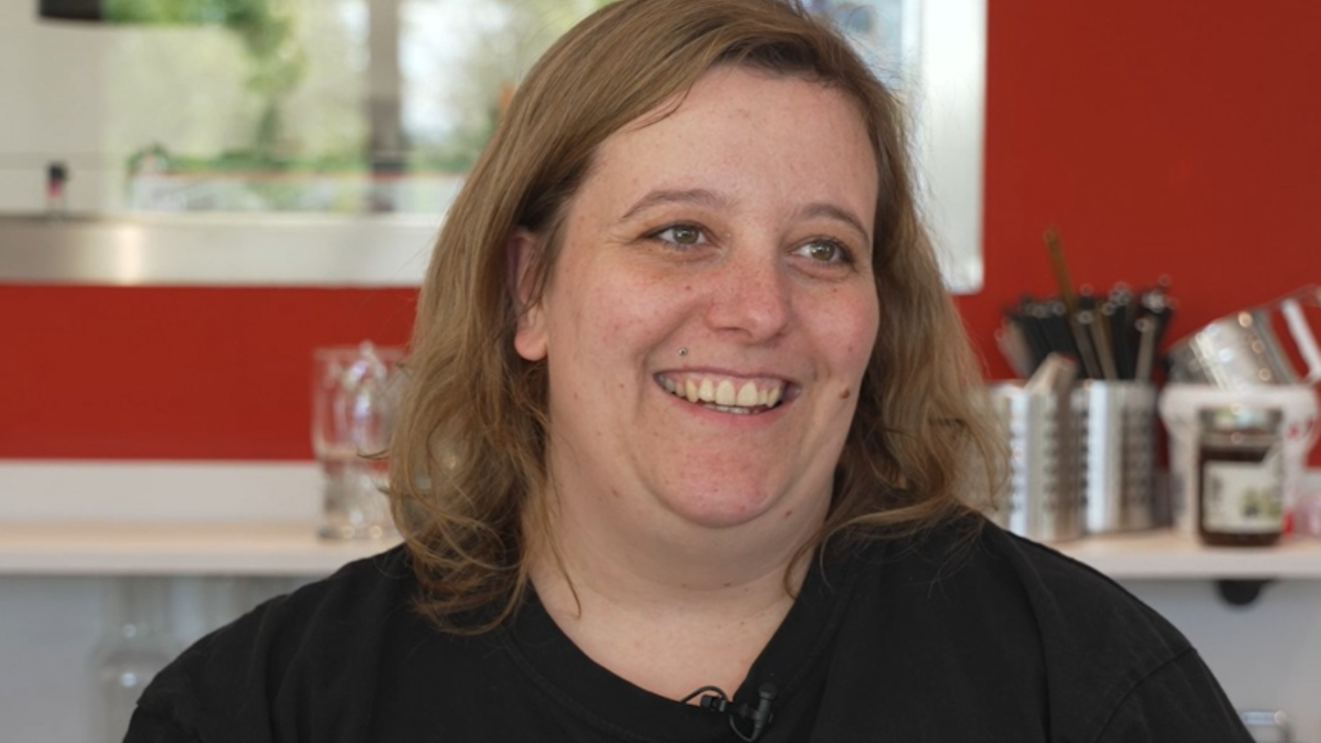 Eine Frau sitzt in einem Innenraum und lächelt, während sie zur Seite blickt. Im Hintergrund sind eine Küche oder Bar mit Gläsern und Utensilien zu sehen.