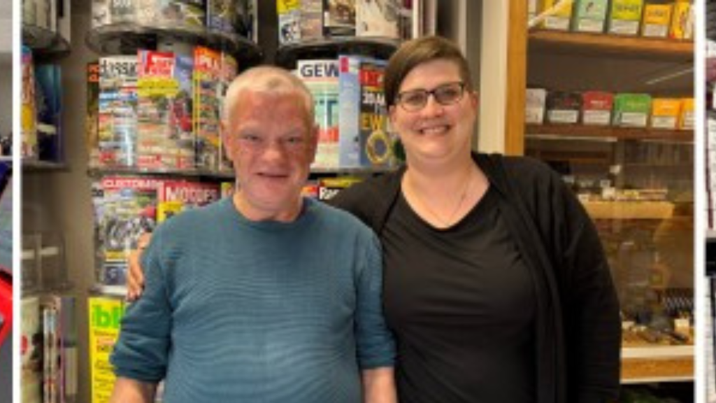 Zwei Personen stehen nebeneinander in einem Geschäft vor einem Regal mit Zeitschriften und lächeln in die Kamera. Eine Person hat den Arm freundschaftlich um die andere gelegt.