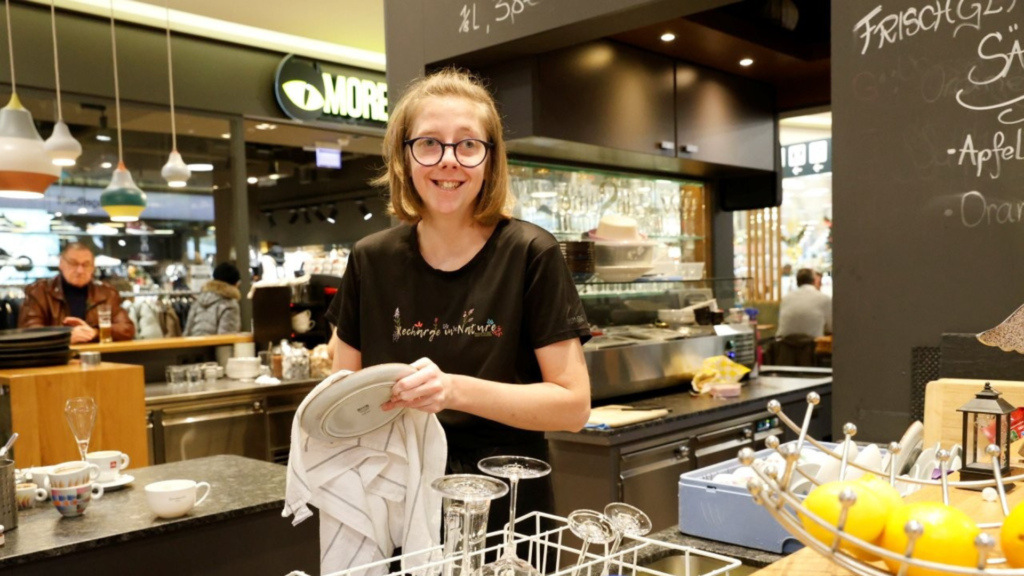 Eine Frau steht hinter der Theke eines Cafés und trocknet Geschirr ab. Vor ihr stehen Gläser und Teller, im Hintergrund sind Kaffeemaschine, Regale und Gäste zu sehen.
