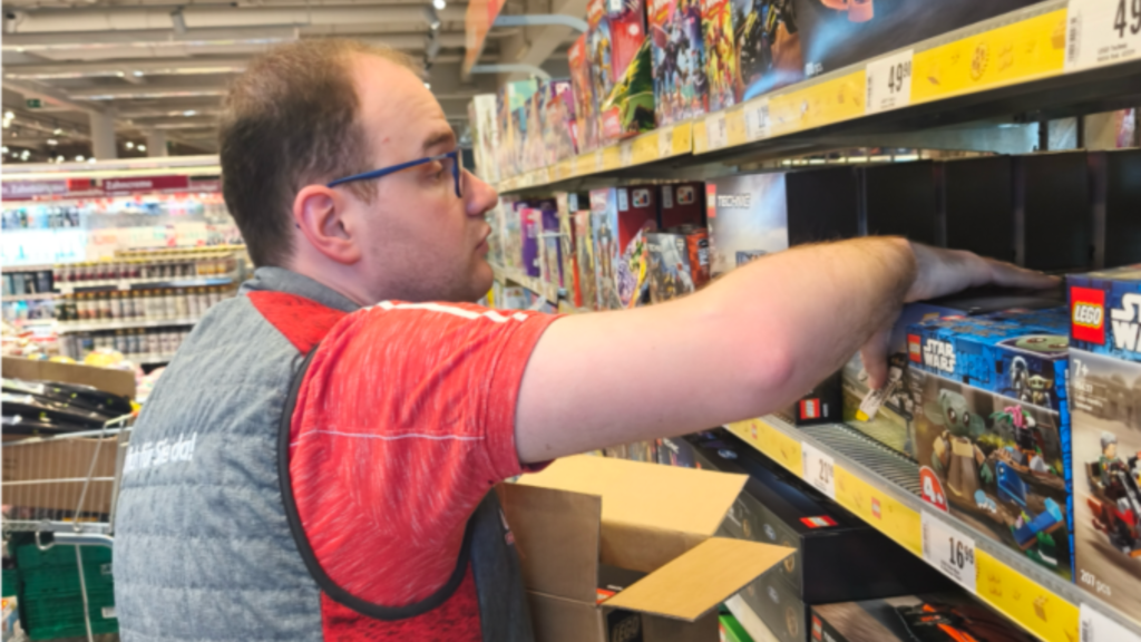 Ein Mann steht in einem Supermarktregal und räumt Spielwaren ein. Er greift nach einer Schachtel und sortiert die Produkte im Regal.