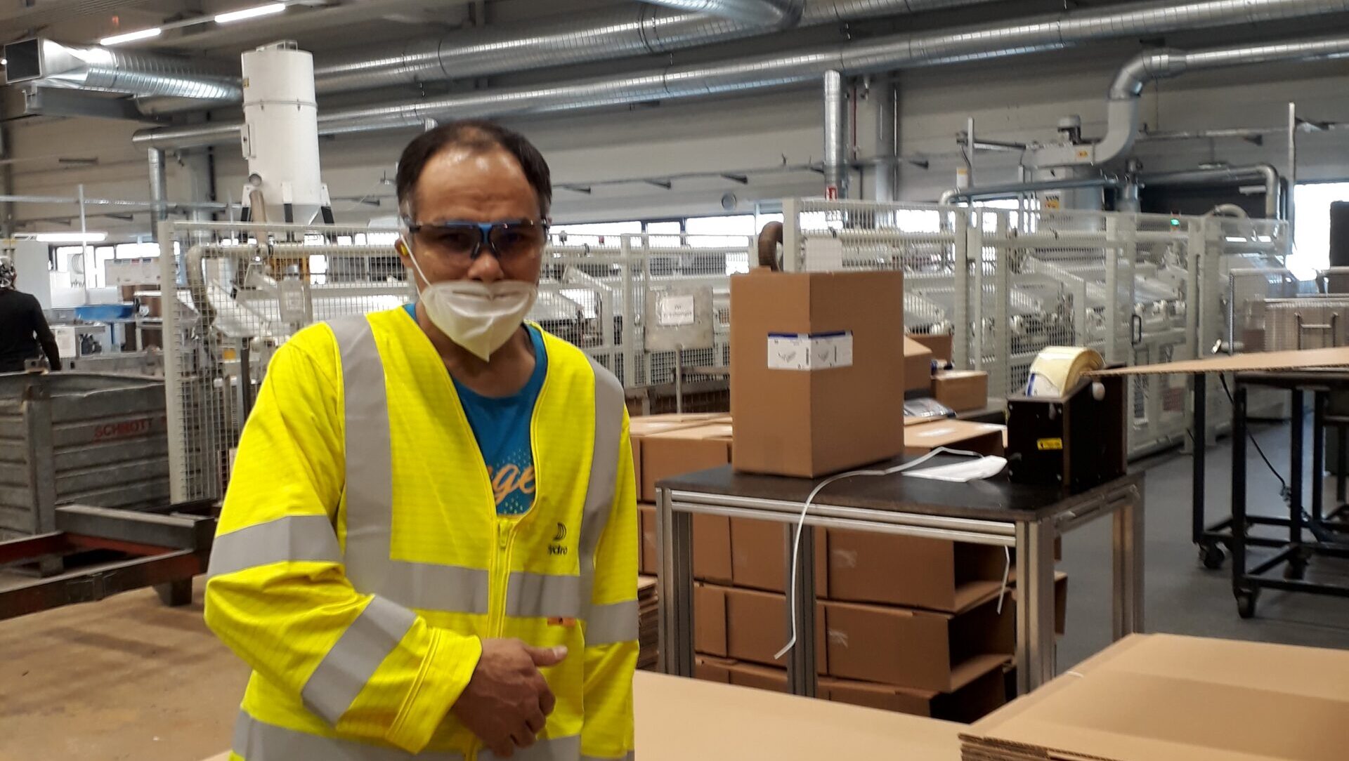 Ein Mann steht in einer Produktionshalle neben gestapelten Kartons und Paletten. Er trägt eine gelbe Warnweste, eine Schutzbrille und eine Maske und lehnt sich an einen Arbeitstisch mit Klebeband und Verpackungsmaterial.
