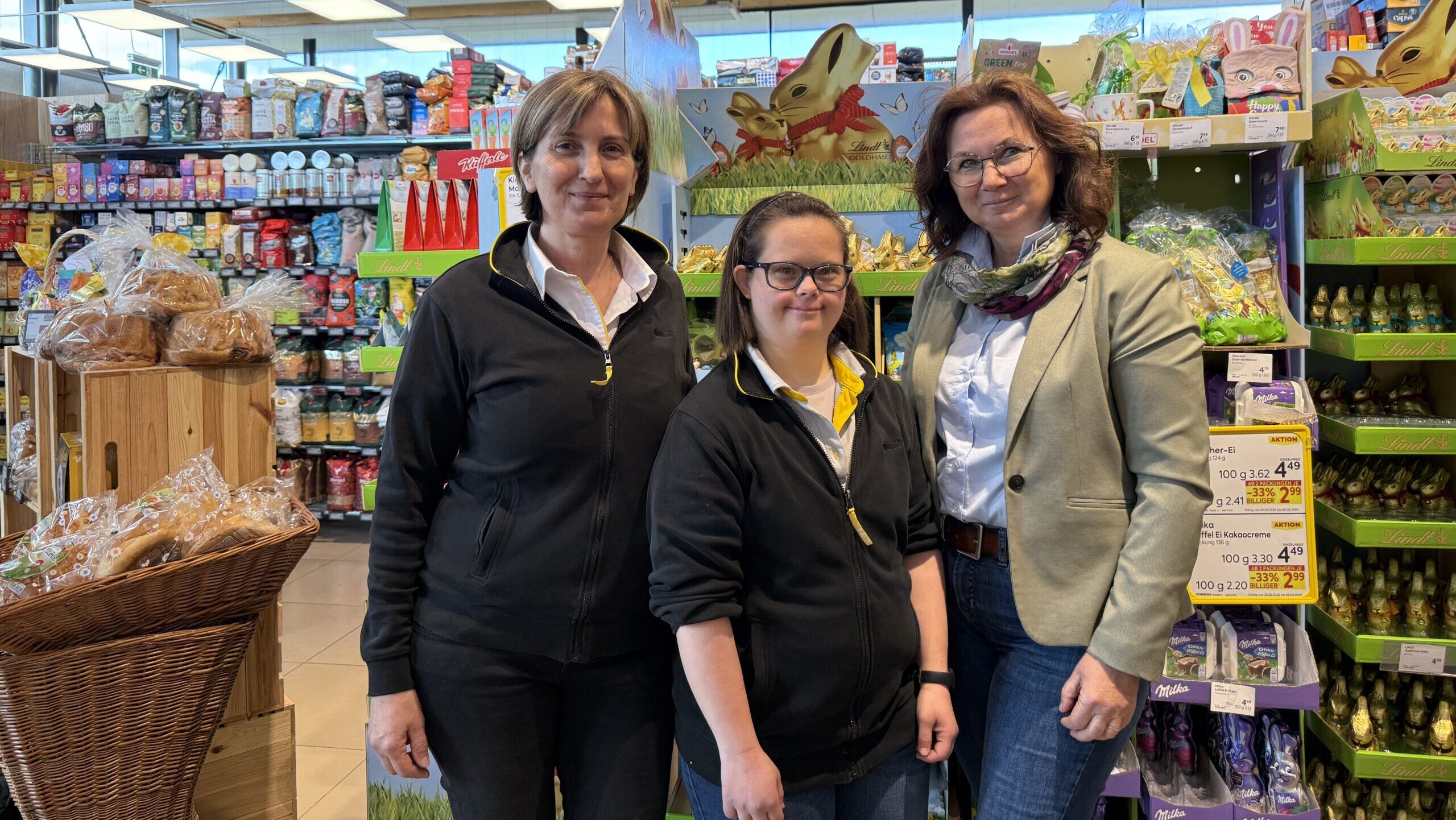 Drei Frauen stehen gemeinsam in einem Supermarkt vor einem Regal mit Waren und lächeln in die Kamera. Hinter ihnen sind gefüllte Regale mit Lebensmitteln und Aktionsaufsteller zu sehen.