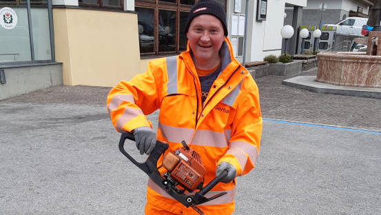 Ein Mann in orangefarbener Arbeitskleidung und Warnjacke steht im Freien und schneidet mit einer motorisierten Heckenschere eine Hecke.