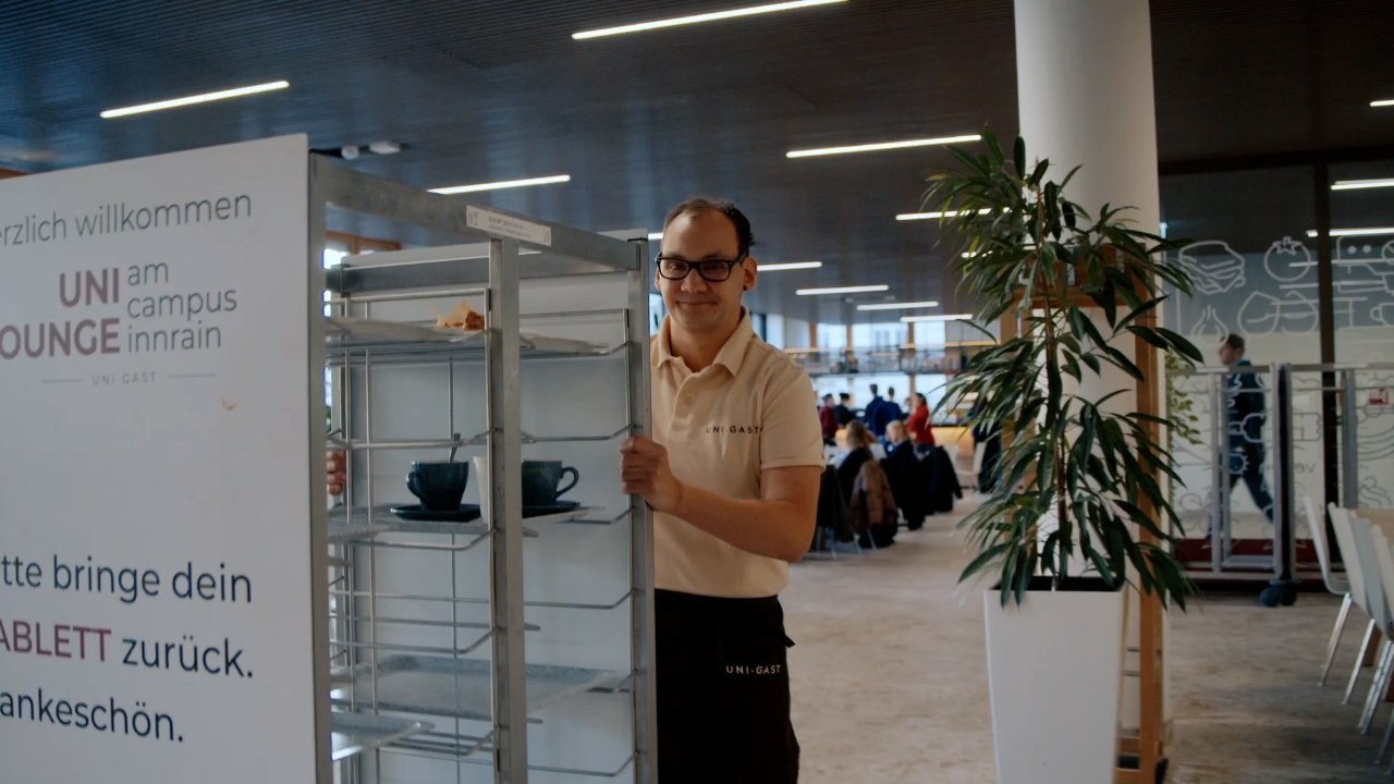 Ein Mann in Arbeitskleidung steht in einer Mensa oder Cafeteria und schiebt einen Servierwagen mit Geschirr. Im Hintergrund sind Sitzbereiche, Gäste und ein großer Innenraum zu sehen.