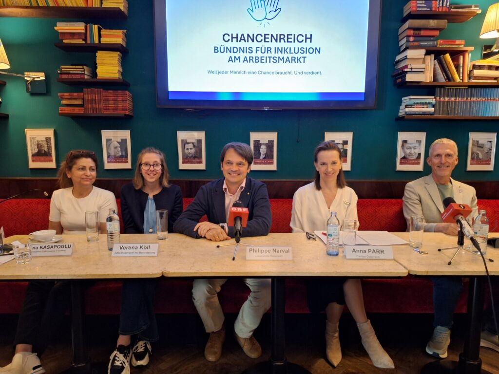 Fünf Personen sitzen an einem Tisch mit Mikrofonen und Namensschildern vor einer Tafel mit der Aufschrift "CHANCENREICH BÜNDNIS FÜR INKLUSION AM ARBEITSMARKT", die auf die Lebenshilfe Österreich - Inklusion und Teilhabe für Menschen mit Behinderungen hinweist. Bücher säumen den Hintergrund.