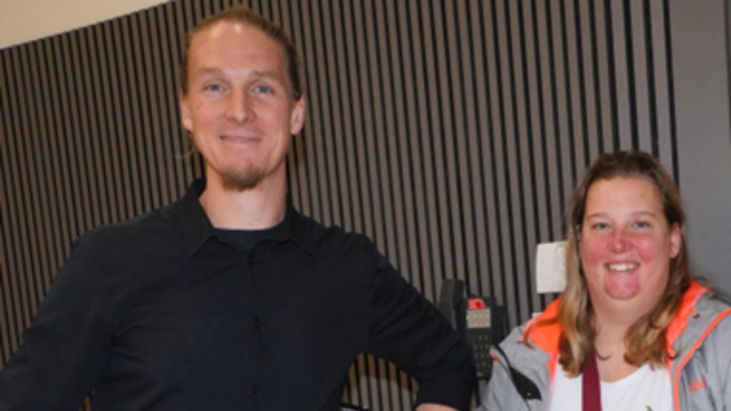 Auf dem Bild sind zwei Personen zu sehen, die nebeneinander stehen und in die Kamera lächeln. * Links: Ein Mann mit zurückgebundenen Haaren und leichtem Bart, trägt ein dunkles Hemd. * Rechts: Eine Frau mit schulterlangen Haaren, trägt eine Jacke mit auffälligem orangefarbenem Innenfutter. Sie stehen vor einer Wand mit vertikalen Holz- oder Lamellenstrukturen, was dem Hintergrund ein modernes, ruhiges Muster gibt. Die Stimmung wirkt freundlich und locker, als wäre das Foto bei einem Treffen oder in einem Arbeitskontext entstanden.