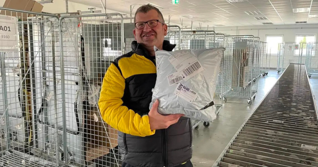 Auf dem Bild sieht man einen Mann in einer Logistik- oder Lagerhalle, der ein Paket in den Armen hält und in die Kamera lächelt. * Er trägt eine schwarz-gelbe Jacke und eine Brille. * In seinen Händen hält er ein graues Versandpaket mit aufgeklebtem Versandetikett. * Im Hintergrund stehen mehrere große Metall-Transportkäfige auf Rollen, wie sie in Verteilzentren verwendet werden. * Rechts im Bild ist ein Förderband zu sehen, das durch die Halle verläuft. Die Umgebung wirkt wie ein Paket- oder Logistikzentrum, und die Szene vermittelt den Eindruck von Arbeit im Versand oder in der Paketverteilung.
