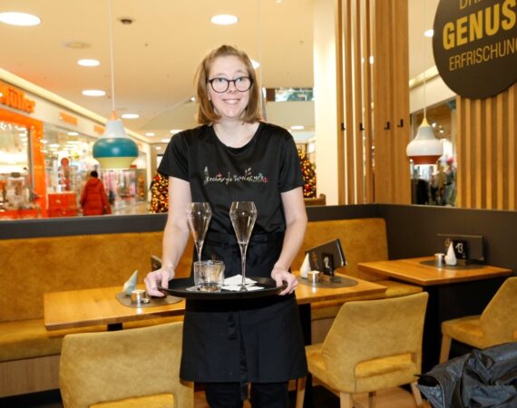 Auf dem Bild sieht man eine junge Frau, die in einem Café oder Restaurant arbeitet und freundlich in die Kamera lächelt. * Sie trägt ein schwarzes T-Shirt, eine Schürze und eine Brille. * In den Händen hält sie ein Tablett mit Gläsern (zwei Sektgläser und ein weiteres Glas). * Sie steht zwischen gedeckten Tischen mit Sitzbänken und Stühlen. * Im Hintergrund erkennt man ein Einkaufszentrum mit Geschäften und Beleuchtung sowie ein rundes Schild mit der Aufschrift „Genuss Erfrischung“. Die Szene wirkt einladend und professionell – sie vermittelt den Eindruck von Servicearbeit im Gastronomiebereich.