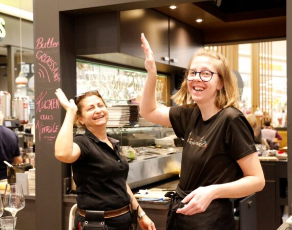 Auf dem Bild sind zwei Frauen in einem Gastronomiebereich zu sehen, die fröhlich miteinander interagieren. * Beide tragen schwarze Arbeitskleidung und stehen hinter einer Theke in einem Café oder Restaurant. * Die Frau rechts (mit Brille) hebt die Hand, ebenso die Frau links – es wirkt wie ein High-Five oder ein gemeinsamer Moment der Freude. * Beide lächeln und schauen in Richtung Kamera, die Stimmung ist locker und positiv. * Im Hintergrund sieht man Küchengeräte, Gläser und eine beschriftete Tafel mit Speisen oder Angeboten. Das Bild vermittelt Teamarbeit, gute Stimmung und Freude an der Arbeit im Servicebereich.