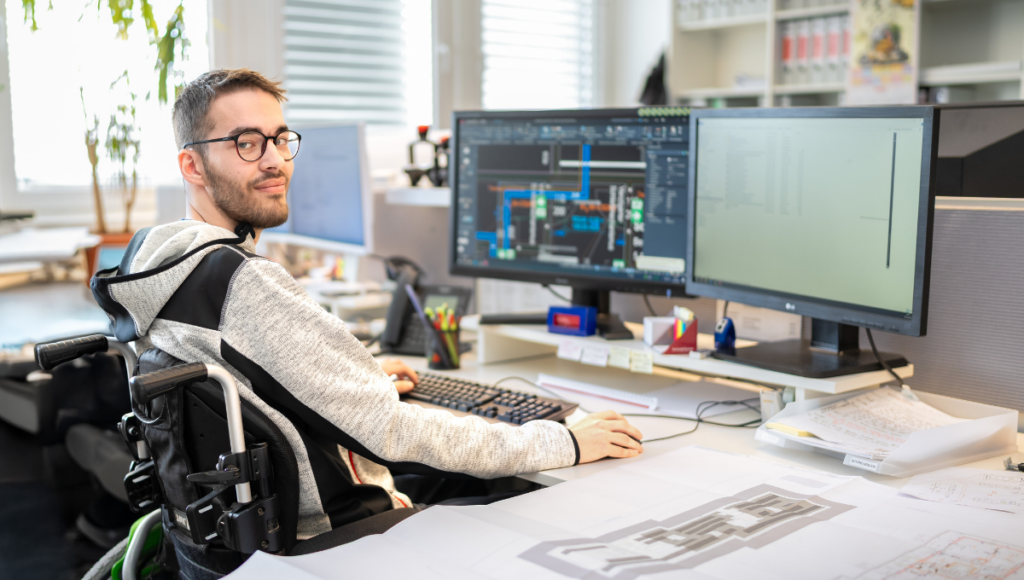 Person sitzt im Rollstuhl an einem Schreibtisch vor mehreren Computerbildschirmen und arbeitet am Computer. Auf den Bildschirmen sind technische Zeichnungen und Programme geöffnet. Auf dem Tisch liegen Pläne und Unterlagen.
