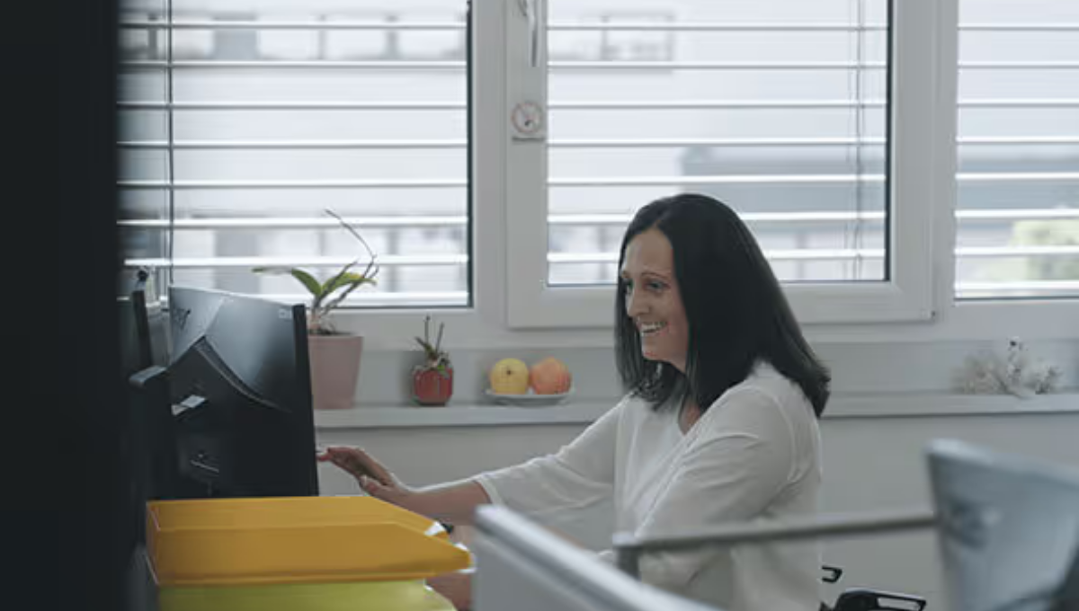 Person sitzt an einem Schreibtisch vor einem Computer und arbeitet. Im Hintergrund sind Fenster sowie kleine Pflanzen auf der Fensterbank zu sehen.