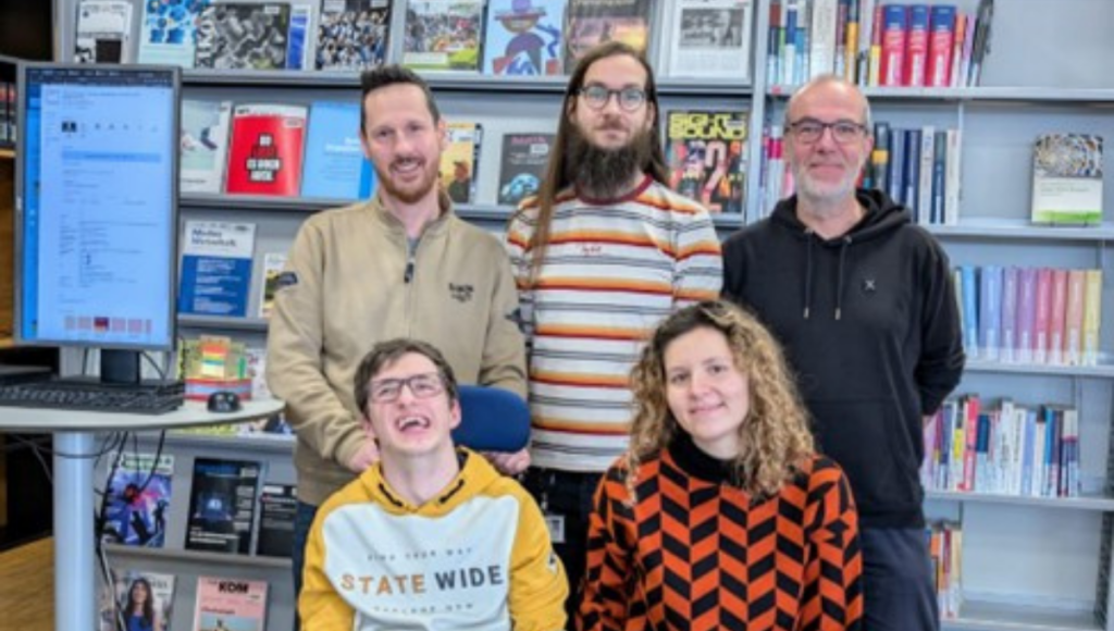 **„Fünf Personen posieren gemeinsam vor Bücherregalen in einer Bibliothek. Eine Person sitzt im Vordergrund in einem Rollstuhl, daneben sitzt eine weitere Person auf einem Hocker. Drei Personen stehen dahinter. Links steht ein Bildschirm mit geöffneter Website.“**