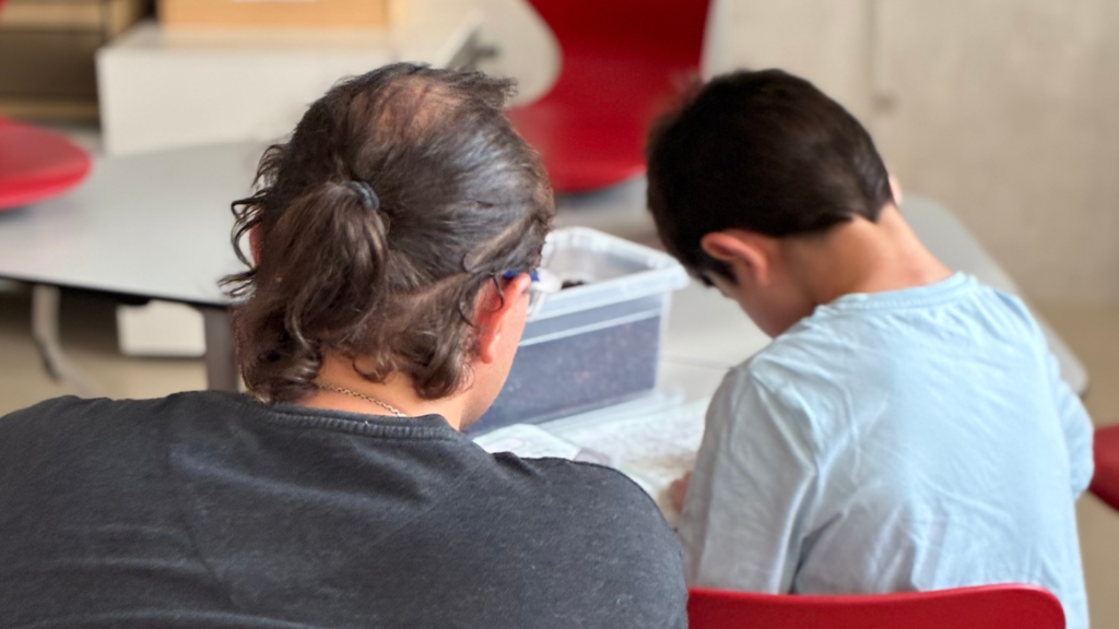 Zwei Personen sitzen nebeneinander an einem Tisch und beschäftigen sich gemeinsam mit Materialien aus einer transparenten Kunststoffbox. Die Aufnahme zeigt die beiden von hinten in einem hellen Raum mit roten Stühlen.