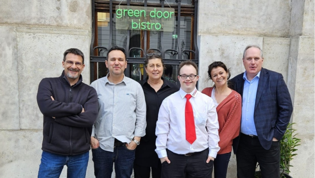 Sechs Personen stehen lächelnd nebeneinander vor dem Green Door Bistro. In der Mitte steht ein Mann mit Down-Syndrom in weißem Hemd und roter Krawatte.