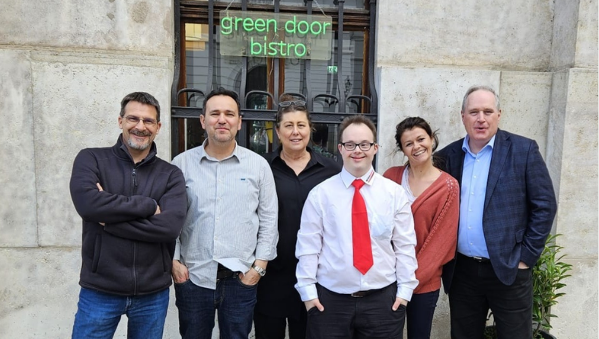 Sechs Personen stehen lächelnd nebeneinander vor dem Green Door Bistro. In der Mitte steht ein Mann mit Down-Syndrom in weißem Hemd und roter Krawatte.